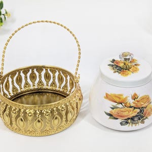 Peut inclure: Un panier en métal doré avec une anse et des détails ornés est placé à côté d'un pot en céramique blanc. Le pot présente un motif floral avec des roses jaunes et un bouton en cristal sur le couvercle. Le panier a une base miroir.