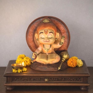 May include: A detailed figurine of a cartoon-like deity with closed eyes, seated and holding a book. The figurine is set against a circular backdrop with script. A small oil lamp, flowers, and a mace are on a wooden base.