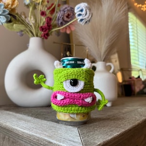 Puede incluir: Un posavasos de monstruo de ganchillo verde con un ojo grande, dientes blancos y boca rosa. El posavasos est&aacute; encima de una lata de refresco. El fondo incluye un jarr&oacute;n de flores y un jarr&oacute;n blanco con una pluma.