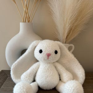 May include: A white crocheted bunny rabbit plush toy with black eyes and a pink nose. The bunny is sitting on a wooden surface.