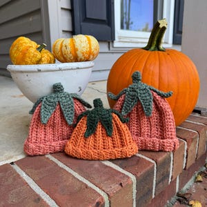 May include: Three crocheted pumpkin hats in shades of orange and pink with green leaf accents sit on a brick surface. A large orange pumpkin and a white bowl with gourds are in the background, suggesting a fall theme.