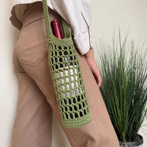 Puede incluir: Un portabotellas de vino verde tejido a ganchillo con una correa larga. El soporte tiene un diseño de red y se ve una botella de vino en su interior. El soporte se lleva sobre el hombro.