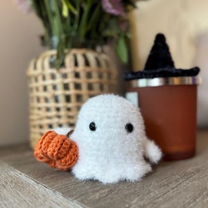 May include: A handmade white ghost plush toy with black button eyes and an orange pumpkin. A black witch's hat sits on a brown candle holder. Flowers are in a vase in the background.