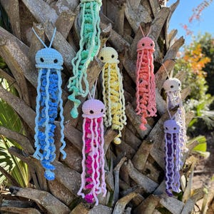 Könnte beinhalten: Eine Sammlung handgefertigter Häkelquallen-Ornamente in verschiedenen Farben, darunter Blau, Grün, Gelb, Rosa und Lila. Jede Qualle hat einen runden Kopf und lange, fließende Tentakel. Sie hängen an einem Baumstamm.