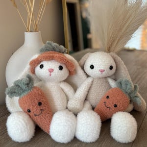 May include: Two white plush bunny rabbits with brown and green carrot toys. The bunnies have fluffy white fur and are sitting on a wooden surface.