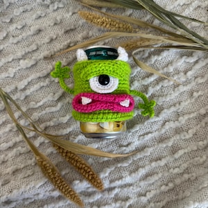 Puede incluir: Un portabotellas de monstruo de ganchillo verde con un gran ojo negro, cuernos blancos y una boca rosa. El portabotellas est&aacute; envuelto alrededor de una lata de refresco. El fondo es una tela texturizada blanquecina con plantas secas.