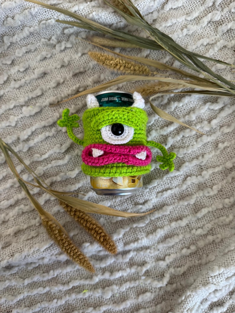 Puede incluir: Un portabotellas de monstruo de ganchillo verde con un gran ojo negro, cuernos blancos y una boca rosa. El portabotellas est&aacute; envuelto alrededor de una lata de refresco. El fondo es una tela texturizada blanquecina con plantas secas.