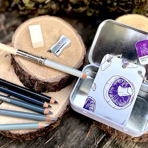 May include: A set of drawing pencils in a tin with a purple mushroom design. The set includes a pencil sharpener, eraser, and a white drawing pencil. The tin has a Pantone colour swatch on the lid.