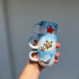 May include: Three stacked ceramic mugs with a sea life theme. The mugs are white with blue accents and feature a red starfish, green turtles, and a grey whale. Each mug has a handle.