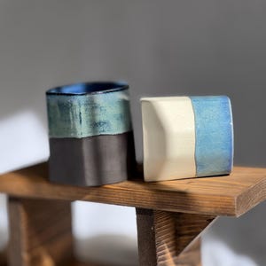 May include: Two ceramic vessels, one cylindrical and one square, displayed on a small wooden shelf. The cylindrical vessel is dark blue and green, the square one is white and blue. The background is a neutral grey.