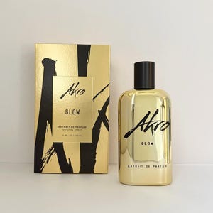 May include: A gold-colored perfume bottle and box set. The bottle and box have the word "Akro" in a black script. The box has black brushstroke designs. The bottle is labeled "Glow" and contains 3.4 fl oz / 100 mL of perfume.