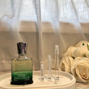 May include: A green and silver perfume bottle with the text "ORIGINAL VETIVER" and three sample spray bottles on a white oval tray. The perfume bottle is 1.7 fl oz and the sample bottles are labeled 2ML, 5ML, and 10ML.