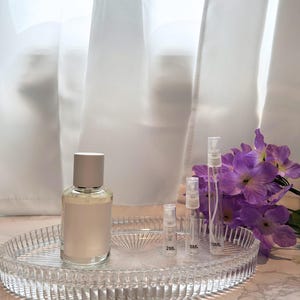 May include: A clear glass perfume bottle with a silver cap and a blank label sits on a clear, oval-shaped glass tray. Several small spray bottles are also on the tray, next to a bouquet of purple flowers. The background is a white curtain.