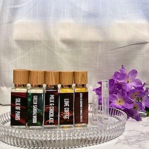 May include: A collection of perfume bottles with wooden caps, each labeled with a different scent: Silk of Paris, Green Sandalwood Wood, Milk & Chocolate, Love Coffee, and Crème de Chestnut & Vanilla Francais. The bottles are on a clear, oval tray, with two sample vials and purple flowers.