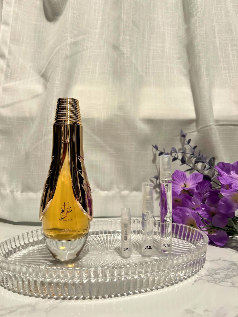 May include: A gold-colored perfume bottle with a decorative design, sitting on a clear, oval-shaped tray. Three clear sample vials are next to the bottle, labeled 2ML, 5ML, and 10ML. A bouquet of purple flowers is in the background.