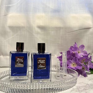 May include: Two blue rectangular perfume bottles with silver caps and labels that read "Flower of Immortality" and "Moonlight in Heaven" by Kilian. The bottles sit on a clear, decorative tray with two small sample vials and purple flowers.