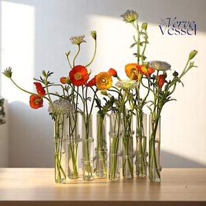 May include: A clear glass vase with multiple cylindrical tubes, each holding a different stem of flowers. The flowers are a mix of orange, red, and yellow, with green stems and leaves. The vase is on a light-coloured wooden surface. The Verve Vessel logo is in the upper right corner.