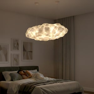 May include: A cloud-shaped pendant light fixture hangs above a bed in a bedroom. The light is white and illuminated, with a textured surface. The bedroom has a neutral color palette, with a bed, curtains, and artwork on the wall.