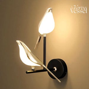 Könnte beinhalten: Moderne Wandleuchte mit zwei vogelförmigen Leuchten. Die Leuchten haben eine goldene Oberfläche und weiße Glühbirnen, die an schwarzen Stielen und einer kreisförmigen Basis befestigt sind. Das Verve Vessel-Logo befindet sich oben rechts.
