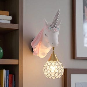 Pode incluir: Arandela de parede com tema de unicórnio. A cabeça branca do unicórnio apresenta um chifre prateado, uma crina rosa e uma coroa floral. Uma luminária dourada suporta uma lâmpada de globo cravejada de cristais, emitindo um brilho quente. A base é preta.