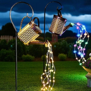 Pode incluir: Duas regadoras decorativas de metal com intrincados padrões recortados estão suspensas em ganchos de pastor de metal preto. As regadoras parecem derramar fluxos de luzes LED brilhantes em um jardim. As luzes são brancas e multicoloridas.