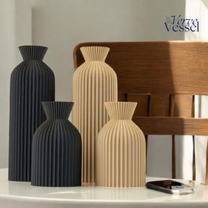 May include: Four decorative vases with a pleated design. Two vases are beige and two are black. The vases are arranged on a white table with a mobile phone in the foreground.