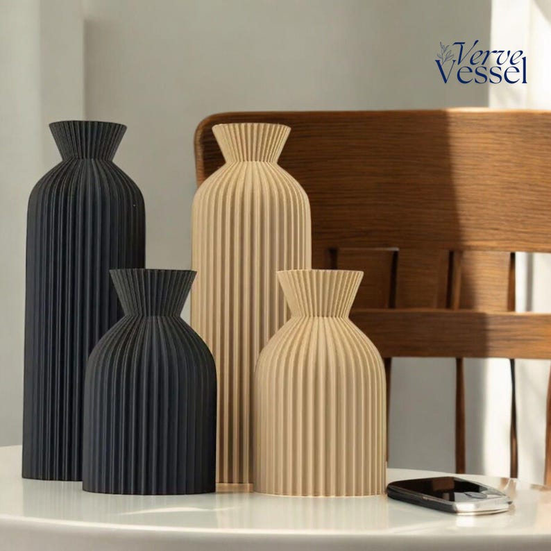 May include: Four decorative vases with a pleated design. Two vases are beige and two are black. The vases are arranged on a white table with a mobile phone in the foreground.