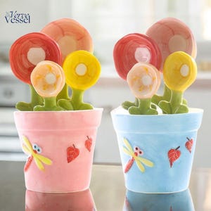 Puede incluir: Dos macetas de cerámica, una rosa y otra azul, cada una con flores decorativas. Las flores son rojas, amarillas y color melocotón. Las macetas tienen diseños de libélulas y hojas. La marca "Verve Vessel" está en la esquina superior izquierda.