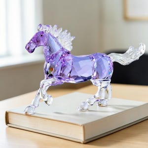 Peut inclure: Figurine de cheval en cristal violet et transparent. Le cheval est représenté au milieu d'une foulée, avec un corps facetté et une crinière et une queue fluides. La figurine est exposée sur un livre.