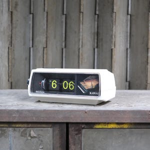 Puede incluir: Un reloj de solapa blanco vintage que muestra las 6:06 en dígitos verde neón. El reloj tiene una esfera negra y la marca "Sanyo" es visible. El reloj está sobre una superficie metálica con aspecto envejecido.