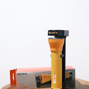 May include: A vintage Sony FL-131 flashlight in yellow and orange, with a black mounting bracket. The flashlight has a textured body and a silver base. The original box is visible, featuring the Sony logo and product details.