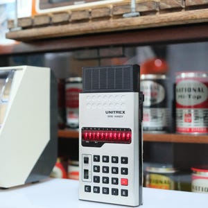 May include: A vintage Unitrex Mini Handy calculator with a silver body, black top, and red LED display. The calculator has black and red buttons. A retro digital clock is visible in the background.