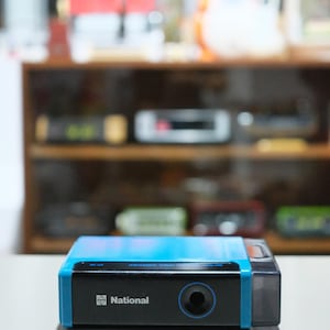 Puede incluir: Un dispositivo de audio National azul y negro. El dispositivo tiene una parte frontal negra con una abertura circular y el nombre de la marca impreso en blanco. La parte superior del dispositivo es azul. El dispositivo está sobre una superficie blanca.