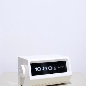 May include: A white retro digital clock displaying 10:00 on a black screen. The clock has a rectangular shape with a dial on the side and the brand name "Sankyo" is visible. The clock is sitting on a light-colored surface.