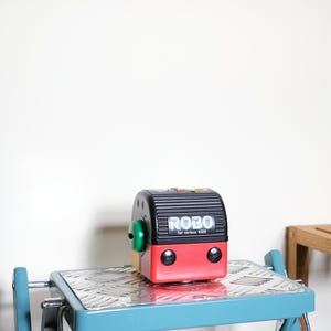 Puede incluir: Un robot de juguete rojo, negro y amarillo con el texto "ROBO for curious KIDS" se muestra en un taburete de metal azul. El robot tiene una perilla verde y dos elementos circulares negros. El taburete tiene una superficie de metal plateado.