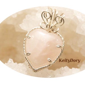 Puede incluir: Un colgante en forma de corazón con una piedra de cuarzo rosa claro, encerrada en un marco de alambre plateado. El colgante tiene un diseño decorativo de alambre en la parte superior. El fondo es una superficie clara y texturizada.