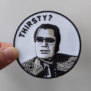 May include: A white embroidered patch with a black and white image of a man wearing sunglasses and a black jacket. The text "THIRSTY?" is written in black above the man's head.