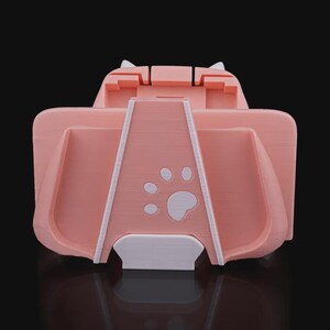 以下が含まれることがあります： 猫の耳のデザインと足跡が付いたピンク色の電話スタンド。スタンドには電話を保持するためのスロットと小さな棚があります。スタンドはプラスチック製で、滑らかな仕上がりです。