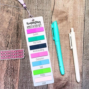 May include: A white rectangular bookmark with a lavender tassel, featuring the text Romantasy Annotation Key and colorful sticky tabs labeled Quotes, Plot Points, World Building, Characters, Sad, Angry, and Love, with a turquoise highlighter and a white pen.