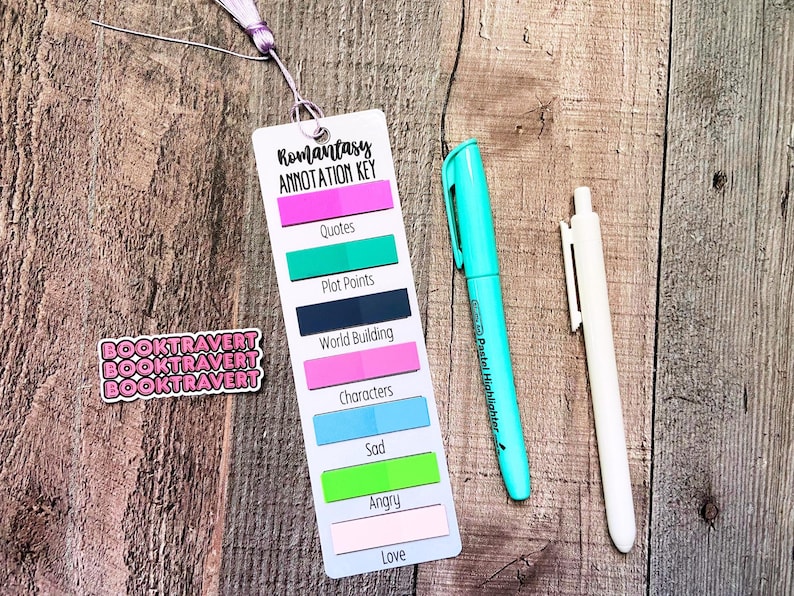 May include: A white rectangular bookmark with a lavender tassel, featuring the text Romantasy Annotation Key and colorful sticky tabs labeled Quotes, Plot Points, World Building, Characters, Sad, Angry, and Love, with a turquoise highlighter and a white pen.
