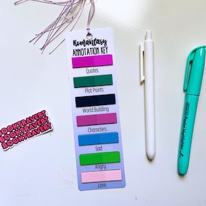 May include: A bookmark with the text "Romantasy Annotation Key" and color-coded sections for annotation. Includes a white pen, a teal highlighter, and a sticker with the text "BOOKTRAVERT" in pink.