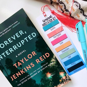 May include: A novel titled "Forever, Interrupted" by Taylor Jenkins Reid, with a bookmark and two pens. The bookmark has a red tassel and labels for annotation: Quotes, Plot Points, Love, Characters, Sad, Angry, and Additional.