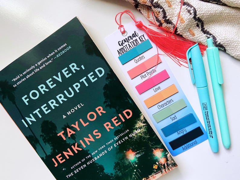 May include: A novel titled "Forever, Interrupted" by Taylor Jenkins Reid, with a bookmark and two pens. The bookmark has a red tassel and labels for annotation: Quotes, Plot Points, Love, Characters, Sad, Angry, and Additional.