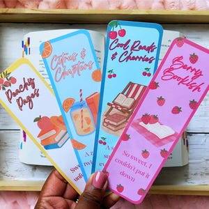 May include: Four colorful bookmarks with fruit and book-themed designs. The bookmarks feature text like "Peachy Pages," "Citrus & Chapters," "Cool Reads & Cherries," and "Berry Bookish." The bookmarks are held over an open book in a wooden tray.