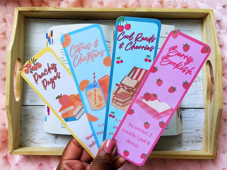 May include: Four colorful bookmarks with fruit and book-themed designs. The bookmarks feature text like "Peachy Pages," "Citrus & Chapters," "Cool Reads & Cherries," and "Berry Bookish." The bookmarks are held over an open book in a wooden tray.