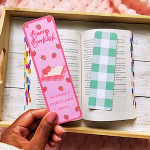 May include: A pink bookmark with the text "Berry Bookish" and strawberry illustrations. The bookmark is held over an open book with a green and white checkered bookmark. The text "So sweet, I couldn't put it down" is also on the bookmark.