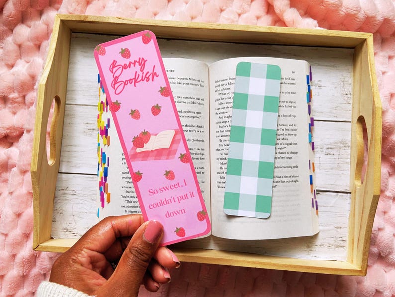 May include: A pink bookmark with the text "Berry Bookish" and strawberry illustrations. The bookmark is held over an open book with a green and white checkered bookmark. The text "So sweet, I couldn't put it down" is also on the bookmark.