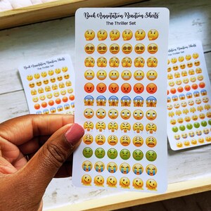 May include: White sticker sheets with various emoji faces in yellow, orange, red, blue, and green. The text "Book Annotation Reaction Sheets" and "The Thriller Set" are printed on the stickers. The stickers are held in a wooden tray.
