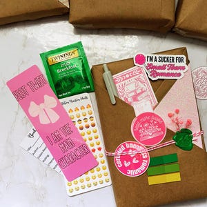 以下が含まれることがあります： 「I'm a sucker for Small Town Romance」と「Certified hopeless romantic」と書かれたピンク色のステッカーが付いた茶色の紙で包まれたパッケージ。 「Plot Twist」と「I am the main character」と書かれたピンク色のブックマークが含まれています。