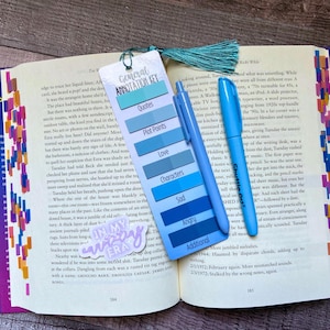 May include: A rectangular bookmark with a teal tassel, featuring a General Annotation Key with color-coded labels for Quotes, Plot Points, Love, Characters, Sad, Angry, and Additional. A blue pen rests on the bookmark, which lies on an open book.